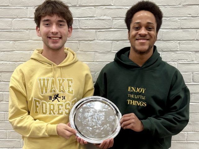 Will and Eleazar standing in front of white brick wall holding silver championship plate