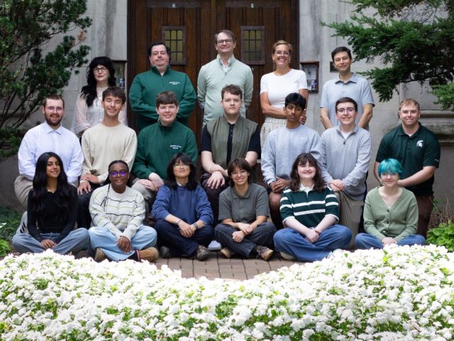 MSU Debate team students and coaches in front of the wooden doors of Beaumont Tower with white flowers in planter in front of them. The staff are pictured standing in the back row and then the debaters are kneeling and seated in front of them. Photo courtesy of Joey Paras, MSU Honors College.