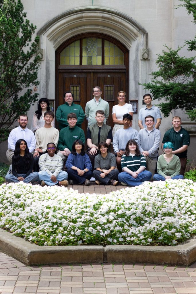 MSU Debate team students and coaches in front of the wooden doors of Beaumont Tower with white flowers in planter in front of them. The staff are pictured standing in the back row and then the debaters are kneeling and seated in front of them. Photo courtesy of Joey Paras, MSU Honors College.